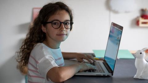Ein Mädchen mit schwarzer ovaler Kunststoffbrille und langen braunen Haaren sitzt vor einem Laptop und sieht aufmerksam in unsere Richtung.