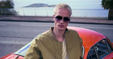 Mann mit Sonnenbrille sitzt auf Auto, hinter ihm das Meer