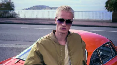 Mann mit Sonnenbrille sitzt auf Auto, hinter ihm das Meer