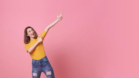 Eine junge Frau mit gelbem T-Shirt und Jeans vor rosa Hintergrund zeigt mit beiden Armen und ausgestreckten Zeigefingern euphorisch nach oben.