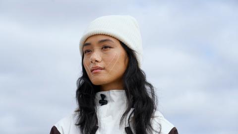 Eine junge Frau mit asiatischen Zügen und weißer Wollmütze und Snowboardjacke hat Gesichtsfeldlinien auf ihrem Gesicht. Es ist eine Frontalaufnahme, ab den Schultern. Ihr Blick geht nach leicht nach rechts.