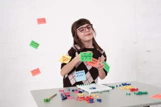 Mädchen mit Brille sitzt am Tisch und hat verschiedene beschriebene Zettel am Pulli