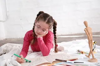 Mädchen mit Brille malt auf dem Boden liegend  in einem Malbuch
