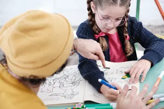 Mädchen mit Brille malt in einem Malbuch, Fotograf zeigt ihr etwas